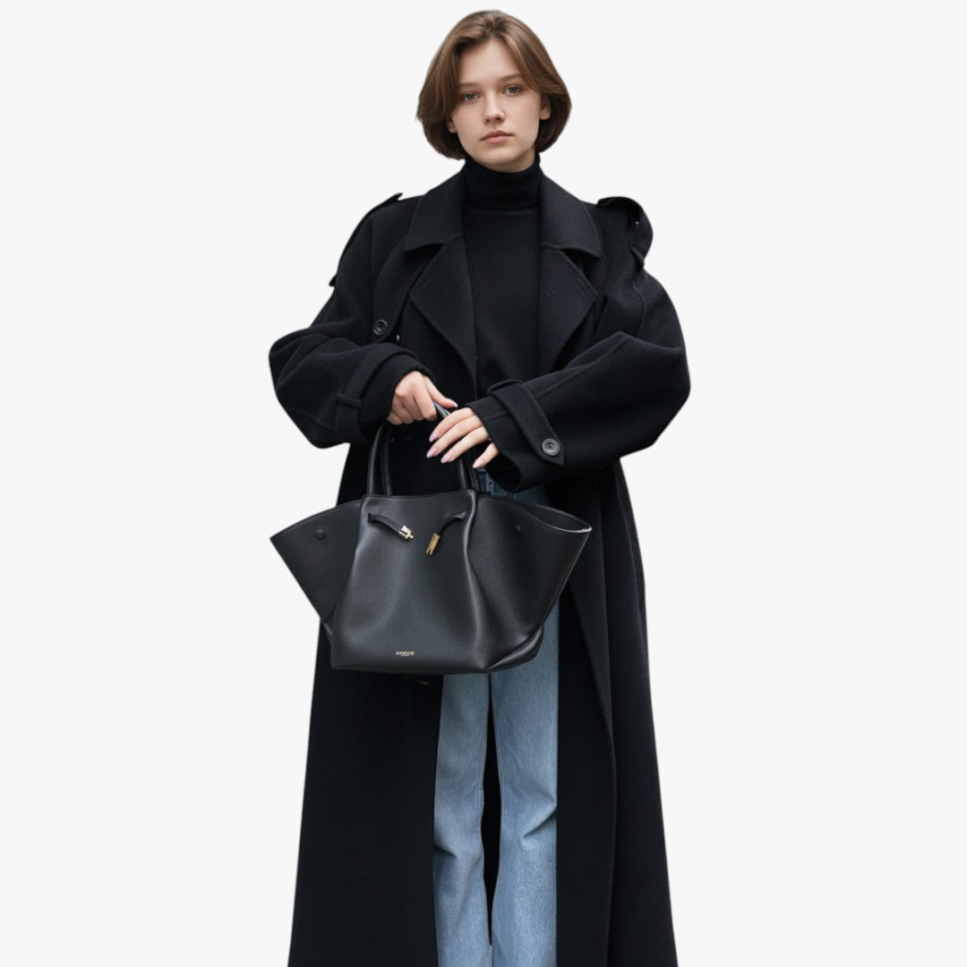 Person wearing a long black coat holding a black handbag on a white background