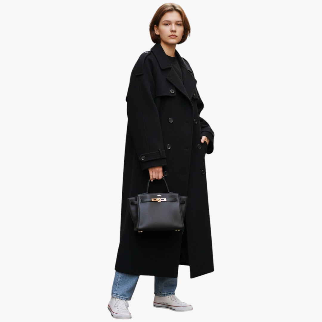 Person wearing a long black coat holding a black handbag on a white background